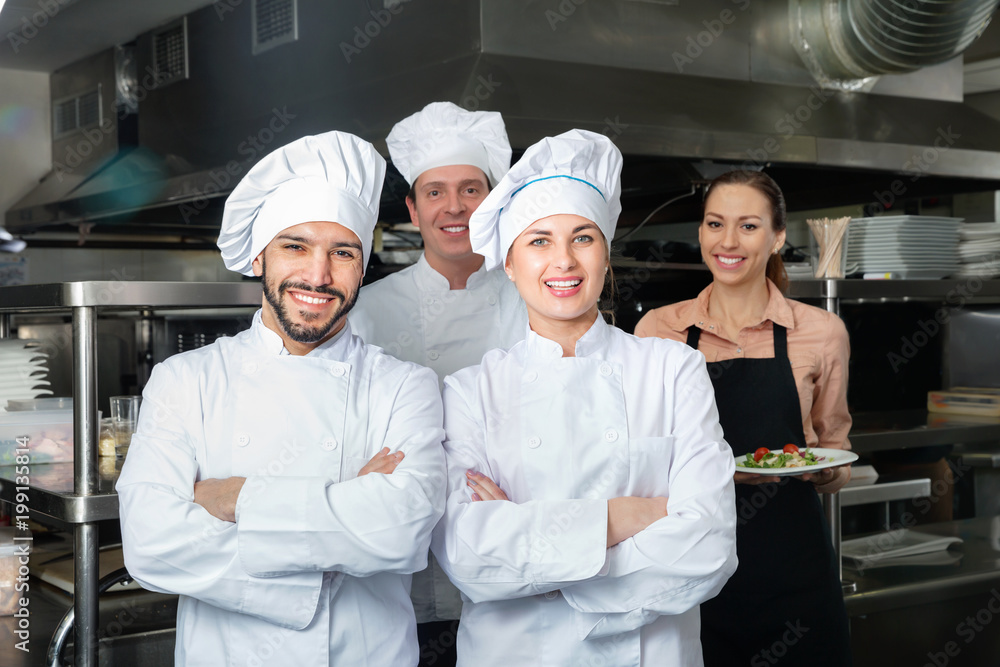 Portrait of two confident cheerful chefs in kitchen with staff of restaurant