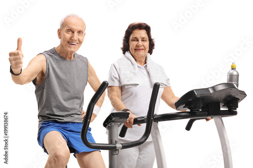Elderly man on an exercise bike making a thumb up sign with an elderly woman on a treadmill