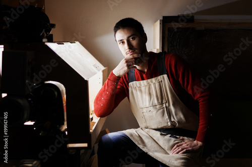 Portrait young male carpenter wearing protective clothes with a beard and a red sweater handles the wood on a lathe in the workshop. dark lighting, carpenters workshop photo