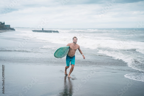 smiling surfer running with surfboard on beach in Bali, Indonesia