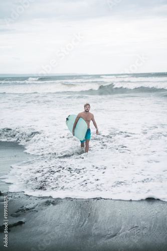 handsome surfer walking from ocean on beach with surfboard in Bali, Indonesia