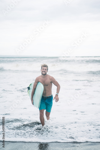handsome smiling surfer running with surfboard in Bali, Indonesia