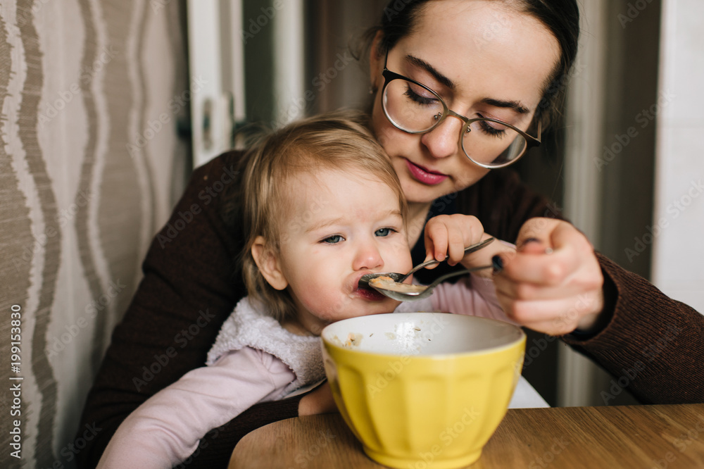 Young happy mother feeding her little baby with delicious tasty soup from plate in kitechen. Funny portrait of eating child. Beautiful girl in glasses together with daughter have diinner at home. Young happy mother feeding her little baby with delicious tasty soup from plate in kitechen. Funny portrait of eating child. Beautiful girl in glasses together with daughter have diinner at home.