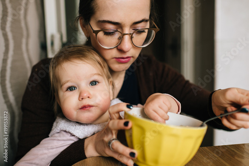 Young happy mother feeding her little baby with delicious tasty soup from plate in kitechen. Funny portrait of eating child. Beautiful girl in glasses together with daughter have diinner at home.