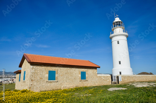 Lighthouse, white lighthouse on blue sky background. Summer, spring, sea and hope concept. Stone building with red roof tiles and blue windows. Green field with yellow flowers. Сyprus, Paphos.