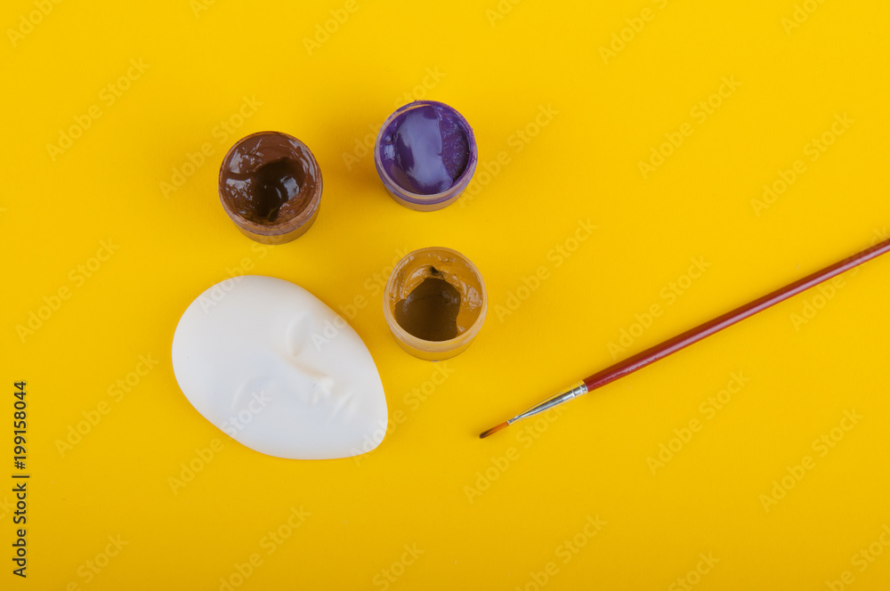 Top view on gypsum mask laying on yellow background near brush, paintings for drawing Top view on gypsum mask laying on yellow background near brush, paintings for drawing
