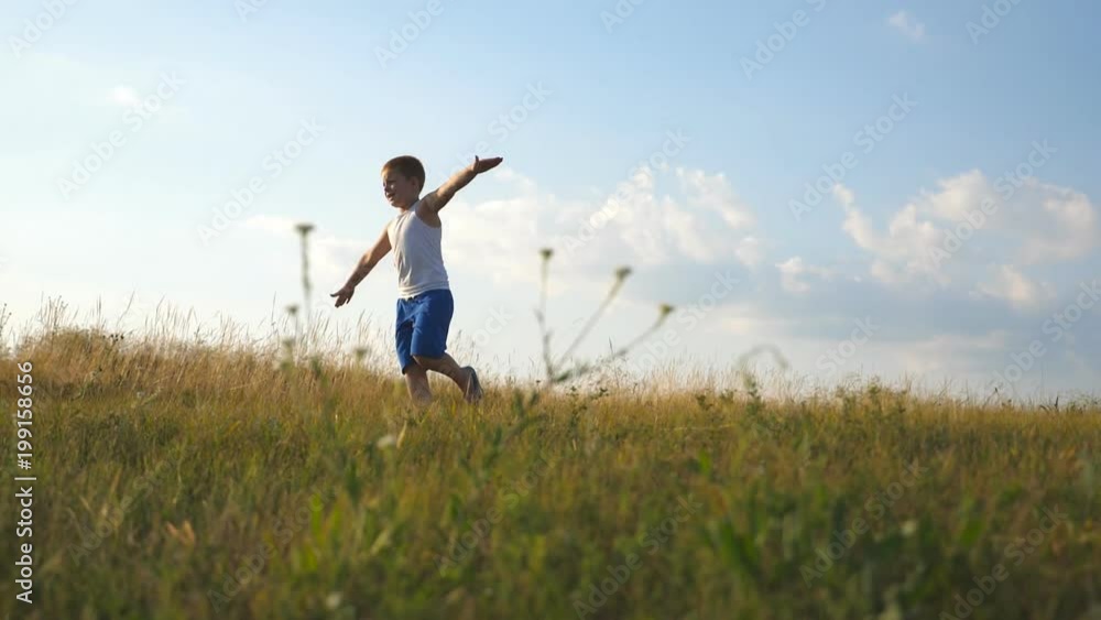 Young boy with raised hands running on green grass at the field on sunny day. Child jogging at the lawn outdoor. Happy smiling male kid having fun in nature on a summer meadow. Slow motion Close up Young boy with raised hands running on green grass at the field on sunny day. Child jogging at the lawn outdoor. Happy smiling male kid having fun in nature on a summer meadow. Slow motion Close up