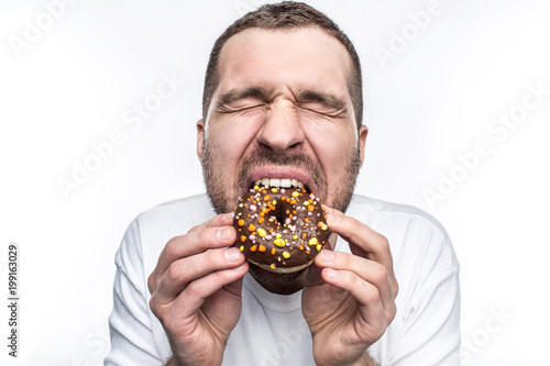 This guy is starving so he is eating a big fat chocolate donut. He has closed his eyes and enjoying the moment. Ypoung man likes to eat different kind of sweets. Isolated on white background.