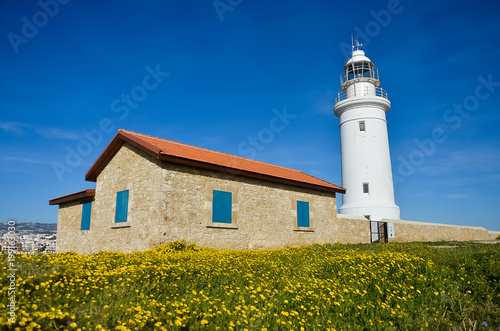 Lighthouse, white lighthouse on blue sky background. Summer, spring, sea and hope concept. Copyspace, place for text. Green field with yellow flowers. Сyprus, Paphos.