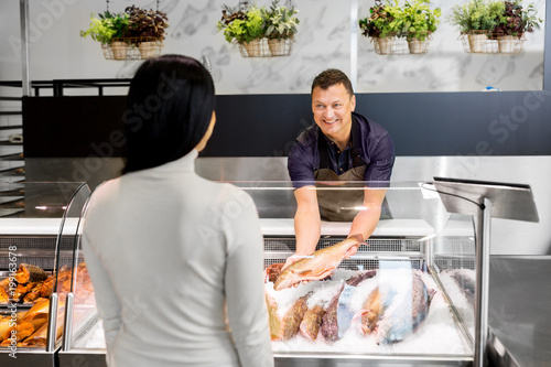 food sale, small business and people concept - male seller showing seafood to female customer at fish shop fridge