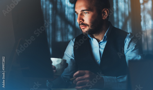 Bearded young businessman working on modern office. Consultant man thinking looking in monitor computer. Manager typing on keyboard in coworking workplace, startup project concept in night with coffee photo