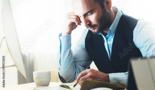Bearded businessman working on modern office. Consultant man thinking looking in monitor computer. Manager writes in notebook in coworking workplace, startup project concept in studio with coffee photo