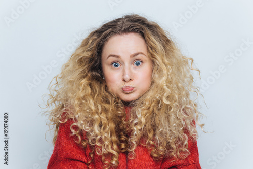 Fuuny blonde female with curly hair, blows cheeks and stares at camera, reacts actively on unexpected news, isolated over white background. Emotional young woman poses indoor. Body language concept © VK Studio