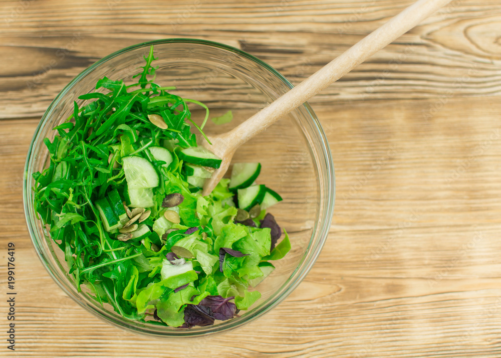 Fresh lettuce leaves, basil, arugula in a glass bowl on a wooden background. Preparation of a vegetarian vegetable salad.