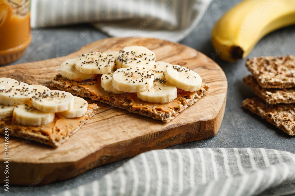 Two multi grain breads with peanut paste, banana slices and chia seeds on a serving wooden board. The concept of healthy fitness breakfast. Two multi grain breads with peanut paste, banana slices and chia seeds on a serving wooden board. The concept of healthy fitness breakfast.
