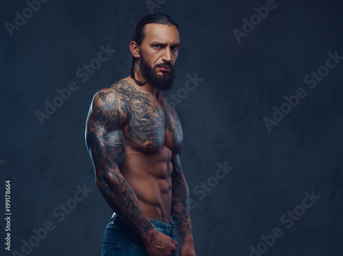 Close-up portrait of a muscular naked bearded tattoed male with a stylish haircut, isolated on a dark background. © Fxquadro