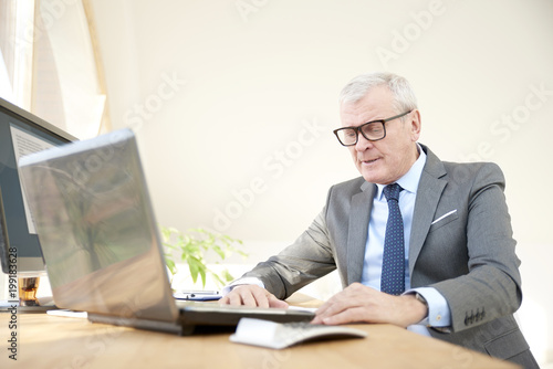 Portrait of an elderly businessman wearing suit while sitting at office desk in front of laptop. 