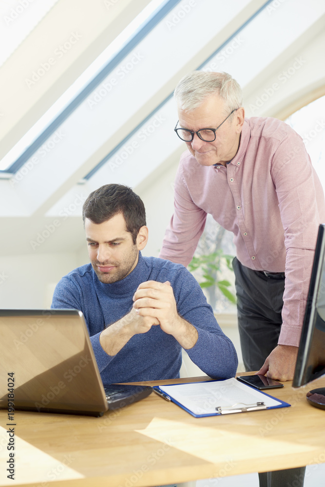Senior businessman giving adice to her young colleague. Sales assistant sitting in front of laptop and consulting with senior manager while working together on new project. Teamwork at the office. Senior businessman giving adice to her young colleague. Sales assistant sitting in front of laptop and consulting with senior manager while working together on new project. Teamwork at the office.