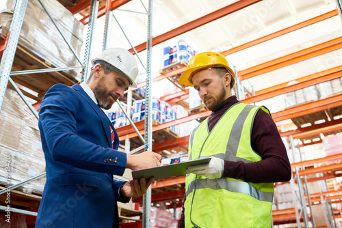 Waist up portrait of mature businessman signing papers on clipboard standing with warehouse worker  discussing inventory