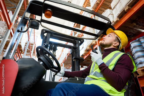 Side view portrait of young man sitting in forklift and using walkie-talkie while moving goods in warehouse, copy space