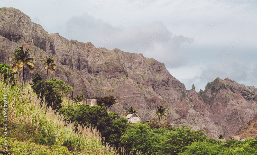 Local stone dwellings surrounded by tropical vegetation. Sharp mountain formation rising on the background. Santo Antao Island, Cape Verde