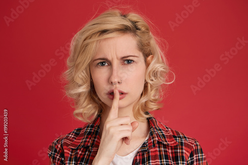 Horizontal shot of young blonde female shows hush sign, please her boyfriend be quite. Isolated over red background photo