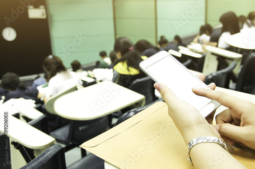 Student/Female teacher using mobile phone in class and blur background of students during study or quiz, test and exams from teacher or in large lecture room / University classroom.