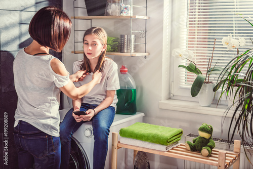 Daughter sitting on washing machine using phone and talking with mother