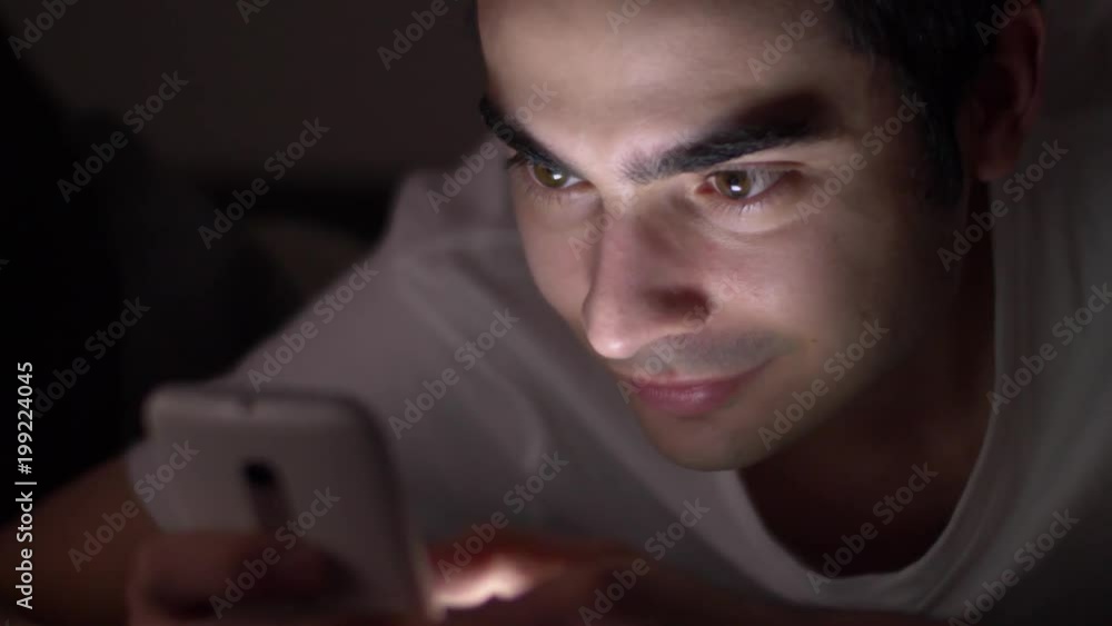 Late at night a man using his smartphone while sitting in his bed in darkness. Late at night a man using his smartphone while sitting in his bed in darkness.