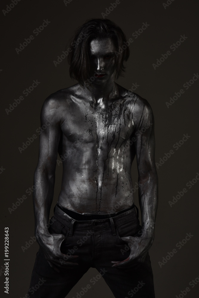 Man with nude torso covered with silver paint, dark background.