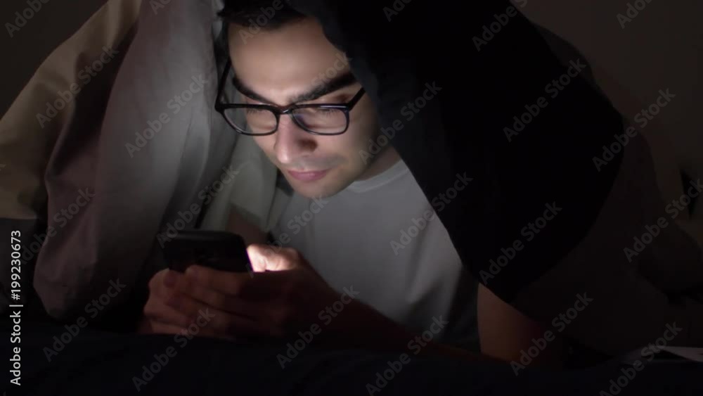 Late at night a man using his smartphone while sitting in his bed in darkness. Late at night a man using his smartphone while sitting in his bed in darkness.