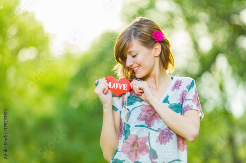 Cute young woman holding a paper heart - shaped sign outdoors.