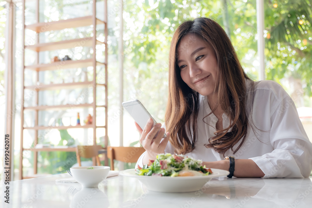 Closeup image of an asian woman using and looking at smartphone with salad in a white plate on table in cafe