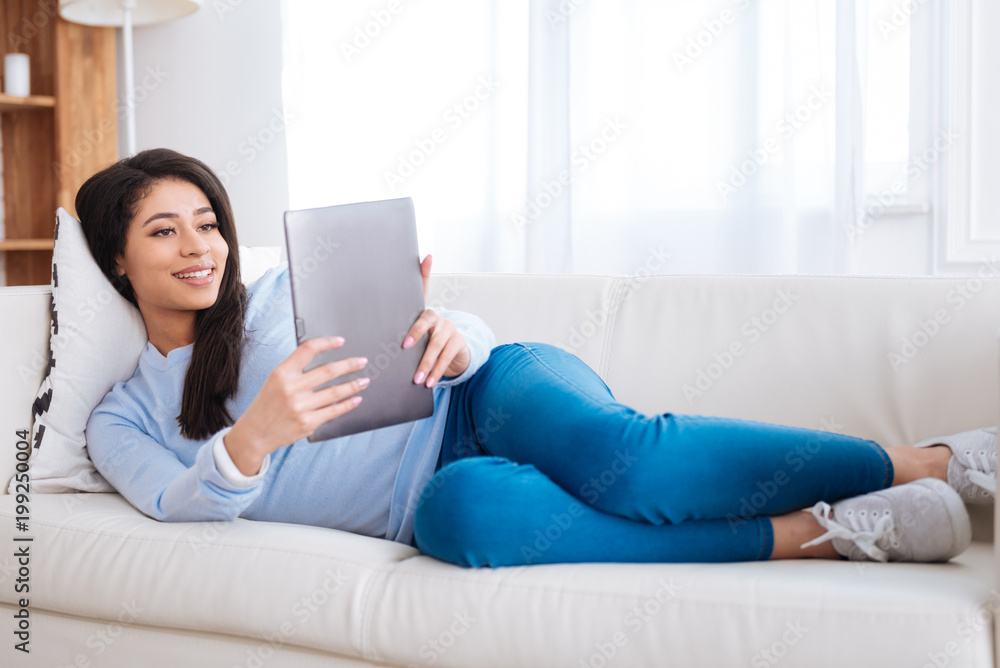 Electronic book. Adorable calm woman rising tablet while resting on couch Electronic book. Adorable calm woman rising tablet while resting on couch
