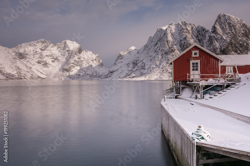 typical Norwegian red cabins on the shore of a snowy fjord with mountains in background
