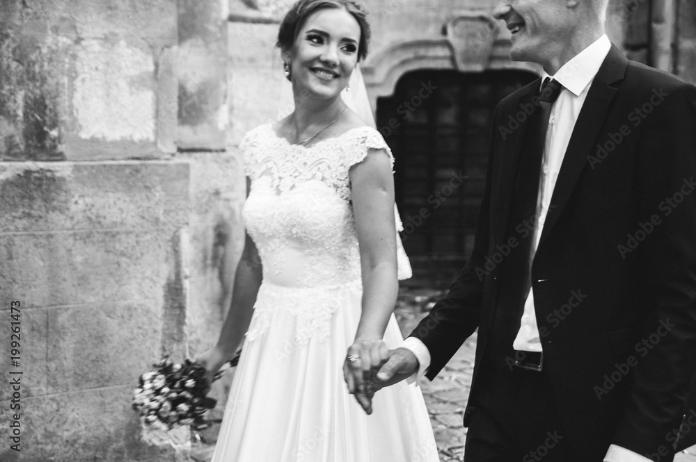 Beautiful wedding couple is walking in the ancient stone walls. Cobblestone road on background. Smiling bride in satin lace dress with veil and happy groom are holding hands and laughing.