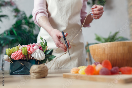 Picture woman of florist with scissor-cutting rope at table with bouquet, marmalade, marshmallow
