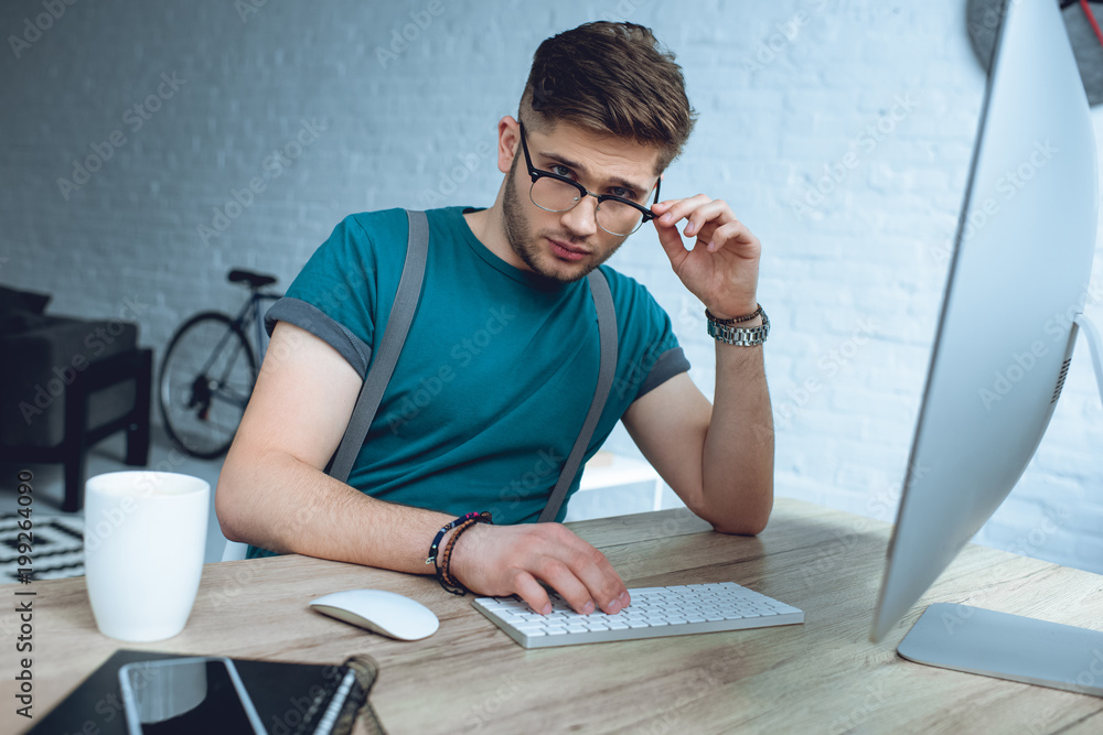 handsome young freelancer adjusting eyeglasses and looking at camera while working with desktop computer