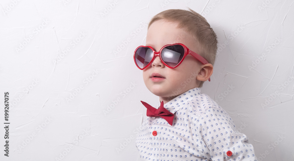 A small serious boy with glasses and a red bow tie is posing against the  white wall. Stock Photo | Adobe Stock