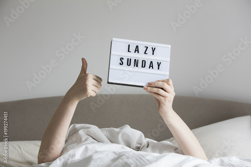 Female in bed under the sheets holding up a lazy sunday sign photo