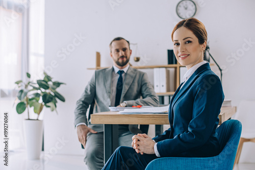 selective focus of smiling businesswoman and colleague behind at workplace in office