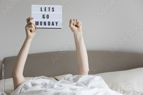 Female in bed under the sheets holding up a lets go monday sign photo