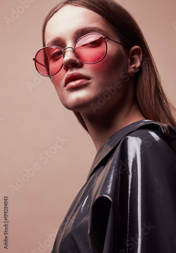 Fashion portrait of beautiful young woman in round red sunglasses and grey latex jacket posing in studio