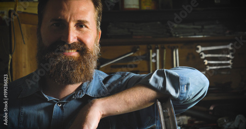 Well-groomed handsome bearded master hipster, specialist in bicycles, repairing a bicycle in his workshop, wheels, frame, spokes, the background of tools. Concept: pro bike, cycle passion, lifestyle.