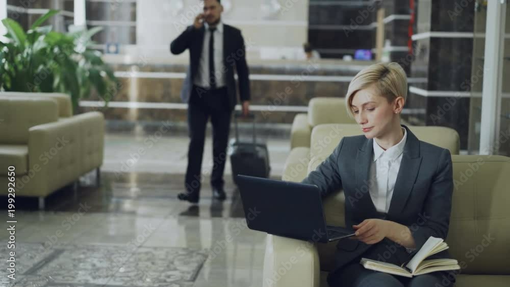 Blonde businesswoman sitting in armchair with notepad and laptop computer while businessman with luggage walking through hotel lobby from reception desk Blonde businesswoman sitting in armchair with notepad and laptop computer while businessman with luggage walking through hotel lobby from reception desk