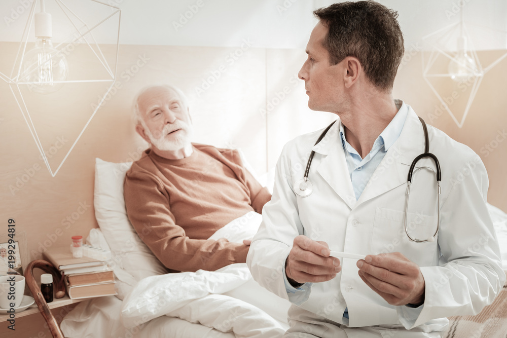 Fototapeta premium What is the diagnosis. Calm skilled handsome medic sitting on the bed holding the thermometer and turning to his patient.