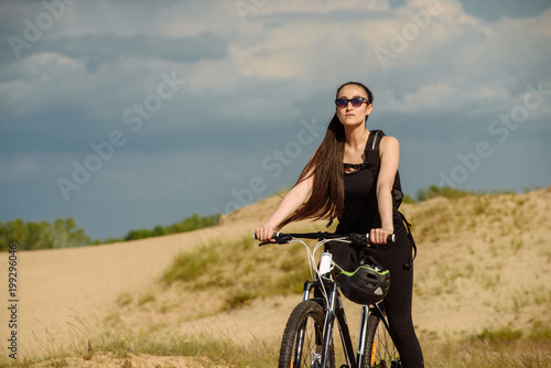 Sport and recreation. Full length image of young female cyclist. Young woman cyclist with backpack rides mountain bicycle on grass field on grey cloud sky