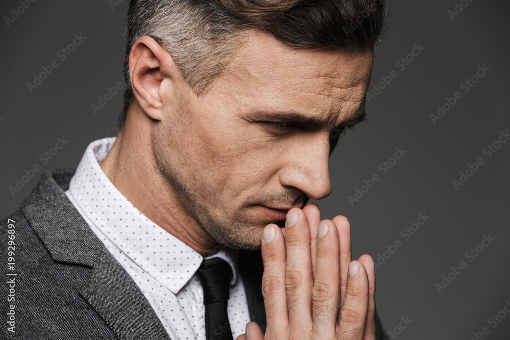 Fototapeta premium Photo closeup of thoughtful calm man wearing classic costume and tie keeping palms together in praying pose, isolated over gray background