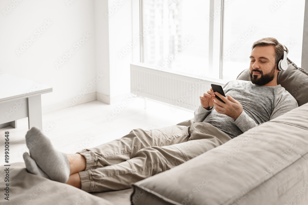 Young guy 30s in basic clothing lying on sofa at home, and listening to music on black mobile phone wearing headphones Young guy 30s in basic clothing lying on sofa at home, and listening to music on black mobile phone wearing headphones