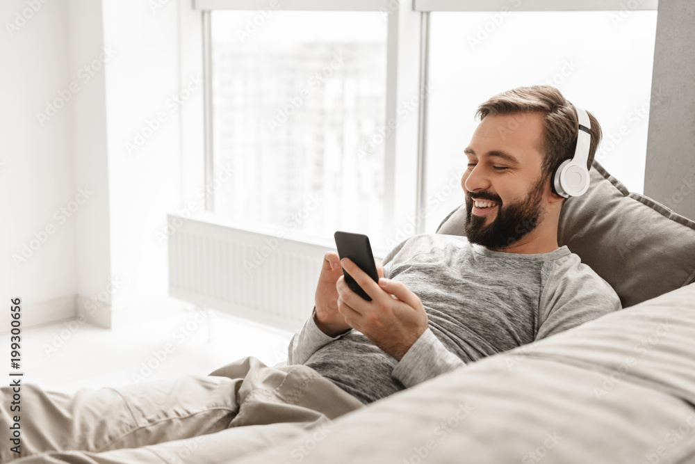 Bearded man 30s in basic clothing lying on sofa at home, and listening to music on black cell phone wearing wireless headphones Bearded man 30s in basic clothing lying on sofa at home, and listening to music on black cell phone wearing wireless headphones